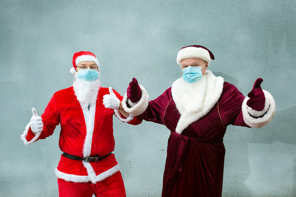 Ded Moroz de Rusia y Santa Claus en máscaras médicas. Las manos de Santa muestran buen signo. Primer plano sobre un fondo gris. - Foto, Imagen