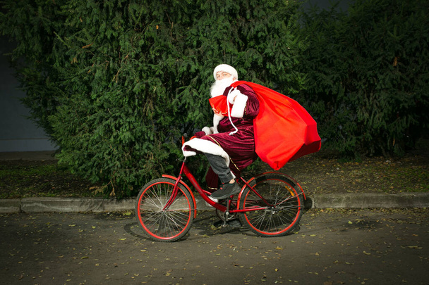 Санта-Клаус ездит на велосипеде и носит с собой большой мешок подарков. Крупный план на фоне хвойных деревьев. - Фото, изображение