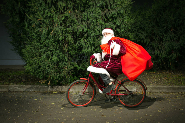 Santa Claus monta en bicicleta y lleva una gran bolsa de regalos. Primer plano sobre el fondo de los árboles de coníferas. - Foto, Imagen