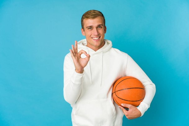Young caucasian man playing basketball isolated on blue background cheerful and confident showing ok gesture. - Photo, Image