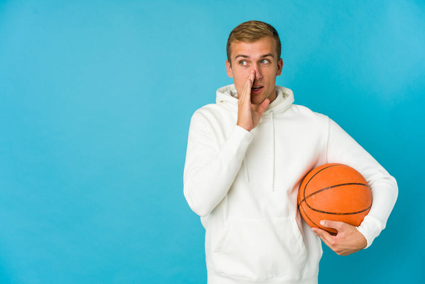 Jonge Kaukasische man die basketbal speelt geïsoleerd op een blauwe achtergrond die het achterhoofd raakt, nadenkt en een keuze maakt. - Foto, afbeelding