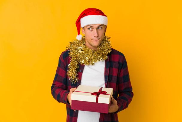Jovem caucasiano com chapéu de Natal segurando um presente isolado no fundo amarelo mostrando um gesto de decepção com indicador. - Foto, Imagem