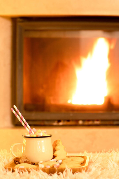 Atmosferyczne zdjęcie z kominkiem na temat świąt Bożego Narodzenia. Piękny kubek tradycyjnego zimowego napoju z piankami, domowe ciasteczka na drewnianej tacy. Koncepcja uroczystości w przytulnym domu. - Zdjęcie, obraz