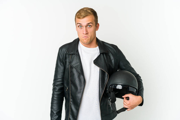 Joven hombre caucásico sosteniendo un casco de moto aislado sobre fondo blanco encoge los hombros y los ojos abiertos confundido. - Foto, Imagen