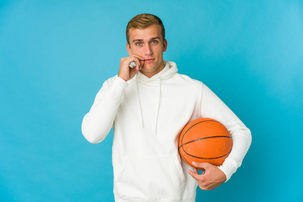 Νεαρός καυκάσιος που παίζει μπάσκετ απομονωμένος σε μπλε φόντο με δάχτυλα στα χείλη κρατώντας ένα μυστικό. - Φωτογραφία, εικόνα