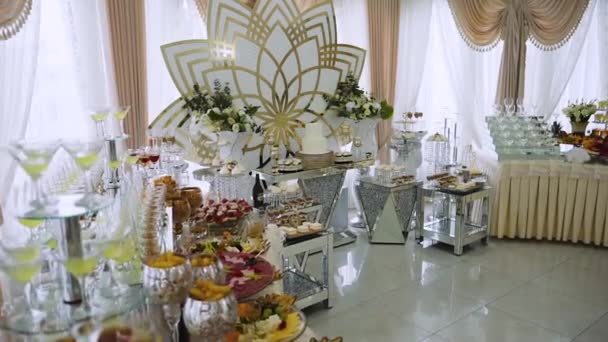 Роскошный дорогой шведский стол с мясом, десертами, фруктами и элитными напитками в бокалах свежих вкусных продуктов находится на блестящих серебряных стендах и на фоне цветка с золотыми элементами в декоре - Кадры, видео