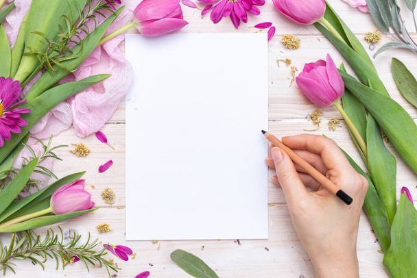 Egy női kéz csúcsképe, amint egy lila virágokkal díszített üres papírra ír. - Fotó, kép
