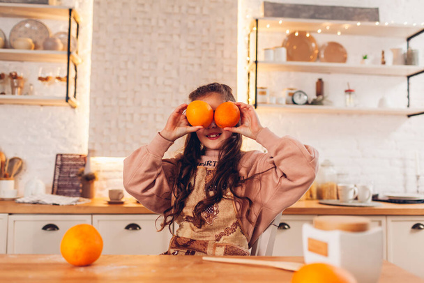 Маленькая девочка играет с двумя фруктами апельсинов вместо глаз на кухне. Ребенок веселится, готовит фартук дома - Фото, изображение