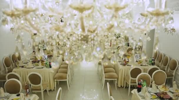 Duża luksusowa restauracja w stylu białym i złotym z zestawem stołów i ozdobione świeżymi kwiatami i numerowanymi cyframi arabskimi, a na suficie żyrandol z wiszącymi błyszczącymi dekoracjami - Materiał filmowy, wideo