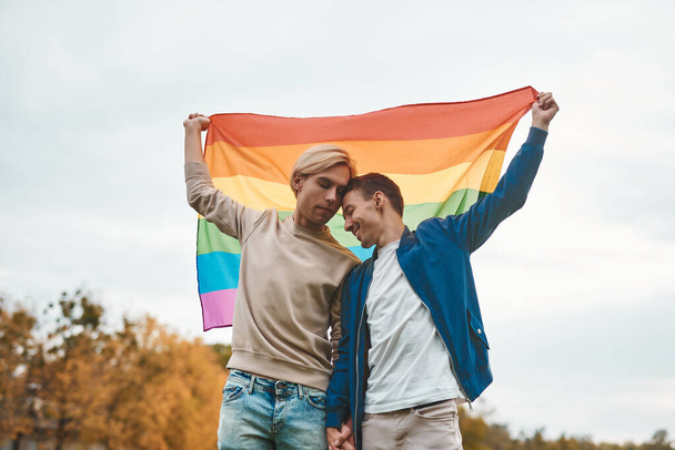 Romantyczna para gejów przytulająca się, całująca i trzymająca się za ręce na zewnątrz. Dwóch przystojniaków z flagą dumy LGBT. - Zdjęcie, obraz