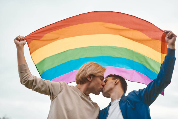 Romantic gay couple hugging, kissing and holding hands outdoors. Two handsome men holding LGBT pride flag. - Photo, Image
