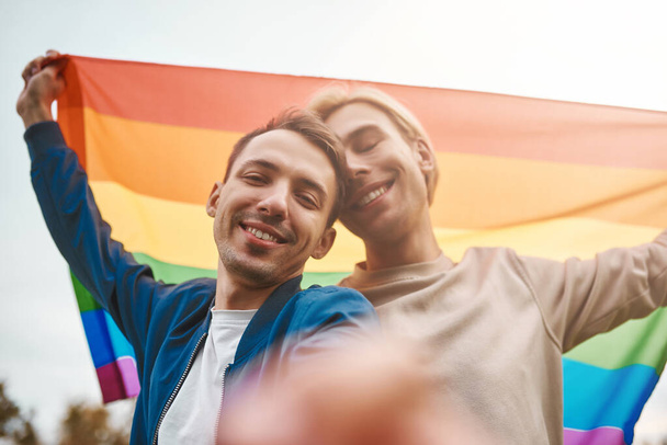 Couple gay romantique étreignant, embrassant et tenant la main à l'extérieur. Deux beaux hommes tenant le drapeau de la fierté LGBT tout en faisant du selfie. - Photo, image