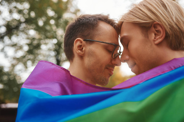 Casal gay romântico abraçando, beijando e segurando as mãos ao ar livre. Dois homens bonitos segurando bandeira do orgulho LGBT no parque. - Foto, Imagem