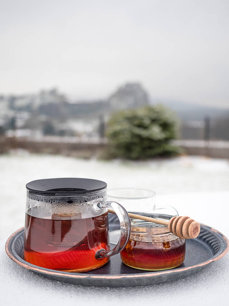 Скляний чайник гарячого чаю і баночка меду на столі, вкритий снігом. Пейзаж на розмитому тлі. Крупним планом з простором для копіювання
. - Фото, зображення