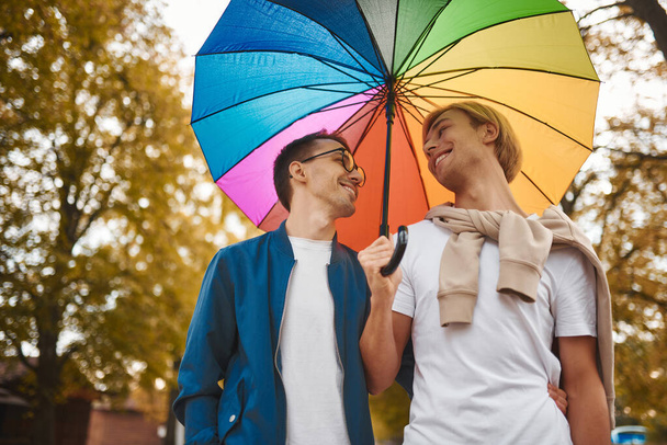 Liebendes schwules Paar, das mit Regenbogenschirm im Freien spaziert. zwei gut aussehende Männer beim romantischen Date im Park. lgbt-Konzept. - Foto, Bild