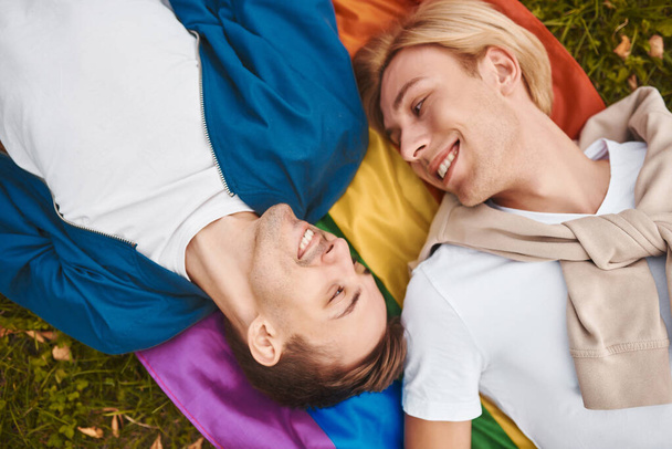 Loving gay couple having romantic date outdoors. Two handsome men lying together on LGBT pride flag. - Photo, Image