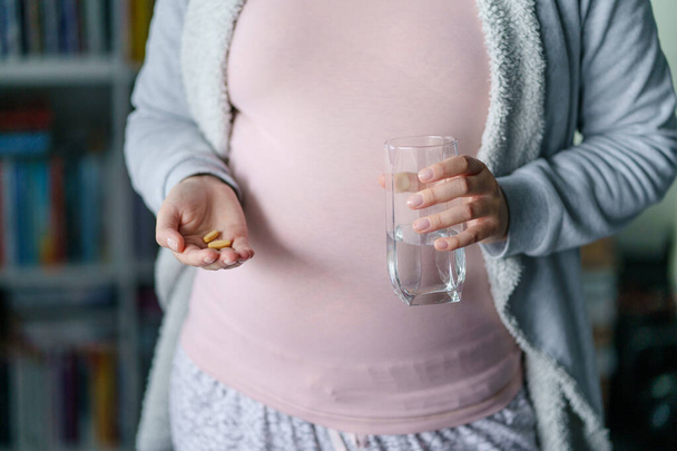 Közelkép középső részén ismeretlen kaukázusi nő terhes anya kezében gyógyszer-kiegészítő gyógyszerek és egy pohár víz - vitaminok szedése a terhesség alatt koncepció - Fotó, kép