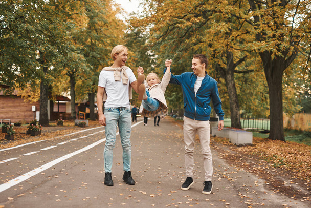 Двое родителей-геев и их приемная дочь вместе гуляют в парке. Счастливая семья ЛГБТ. - Фото, изображение