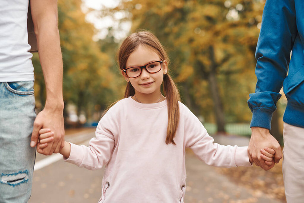 Vágott kép két meleg szülőről, akiknek az örökbefogadott lányuk fogja egymás kezét, és együtt sétálnak a parkban. Boldog LMBT családi koncepció. - Fotó, kép