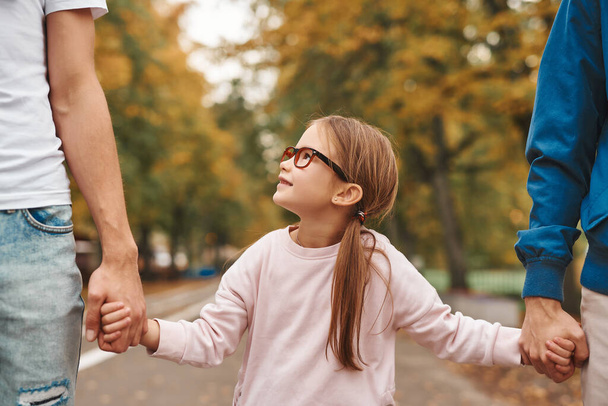 Das Bild zeigt zwei schwule Eltern mit ihrer Adoptivtochter, die sich an den Händen halten und gemeinsam im Park spazieren gehen. glücklich lgbt familienkonzept. - Foto, Bild