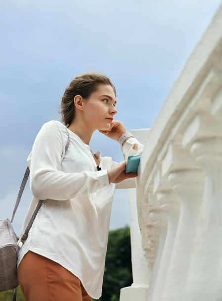 Young woman in white blouse stands near concrete railing and stares into the distance outdoors, in vertical format with copy space - Фото, изображение