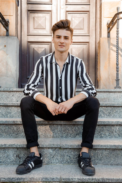 Young man sitting on the stairs in front of the house. Stylish guy in stylish clothes. Young Italian man on the street of the old town. Guy model posing on the stairs. - Foto, Imagen