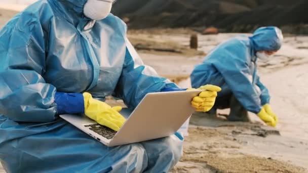 Medium close-up van vrouwelijke ecologie specialist in beschermende kleding en ademhalingsmasker werken op laptop verzamelen van gegevens uit vervuild water gebied met collega - Video