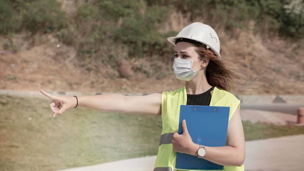 Jovem trabalhadora da construção com capacete branco e máscara facial apontando com a mão no canteiro de obras. - Foto, Imagem