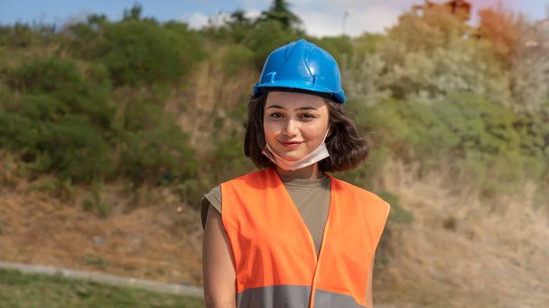 Ritratto di una bella giovane donna allegra ingegnere o operaio in un cappello rigido blu uniforme e protettivo, sorridente e guardando la macchina fotografica in cantiere. - Foto, immagini