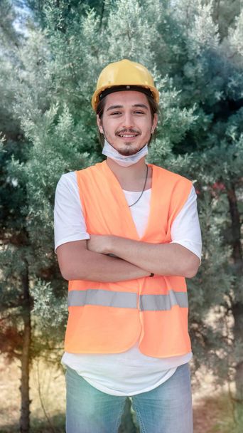 Porträt eines jungen männlichen Bauarbeiters mit gelbem Helm und Mundschutz, der mit verschränkten Armen auf der Baustelle steht und in die Kamera blickt. - Foto, Bild