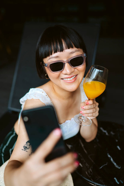 Asiatico capelli corti donna indossare occhiali da sole prendere selfie con succo d'arancia. - Foto, immagini
