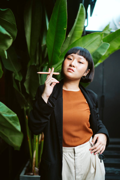 Retrato de joven asiática mujer pelo corto celebración pluma como cigarrillo en la naturaleza. - Foto, Imagen