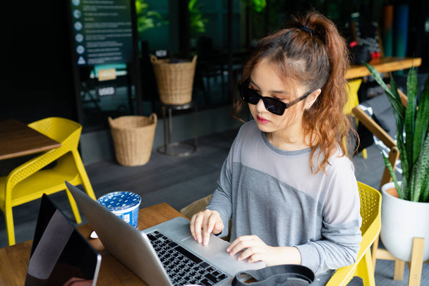 美しいアジアの女性は、屋外のカフェでラップトップで働いてサングラスをかけて. - 写真・画像