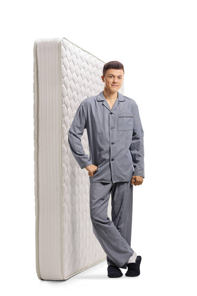Full length portrait of a young man in pajamas leaning on a bed mattress and smiling isolated on white background - 写真・画像