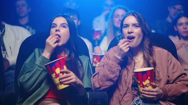 Счастливые женщины едят попкорн в кинотеатре. Весёлые девушки смотрят кино. - Фото, изображение