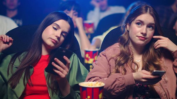 Zaklatott nők ellenőrzik a mobilt a moziban. Lányok görgetés okostelefon beltéri. - Fotó, kép