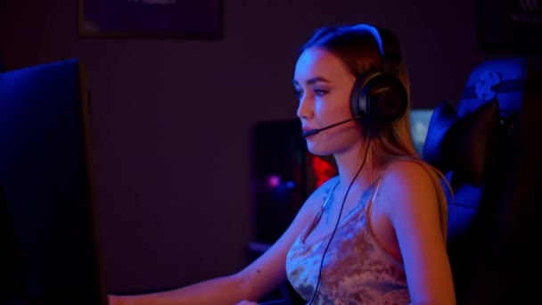 Tattooed gamer girl in big headphones sitting by the PC in the neon gaming club and playing online game - Footage, Video