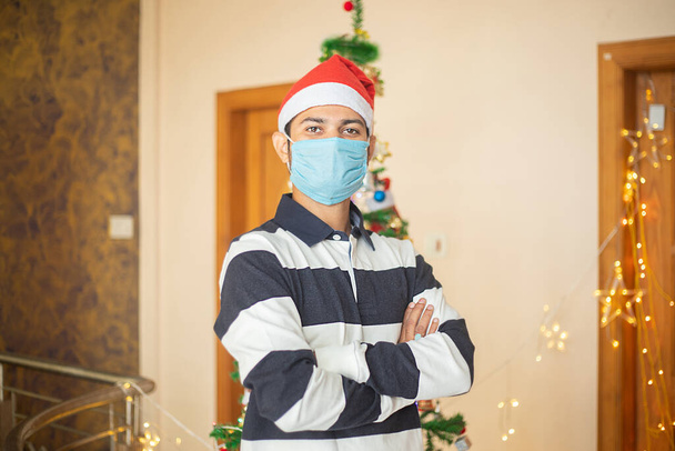 Портрет молодого чоловіка в масці та капелюсі Санти, що стоїть на хресті, концепція святкування Різдва, пандемія ковадла-19
. - Фото, зображення
