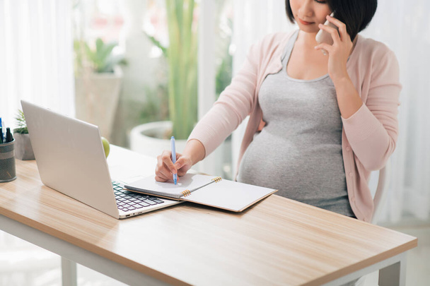 Zwangere zakenvrouw werken met laptop en praten over de telefoon in gezellige heldere kantoor. - Foto, afbeelding