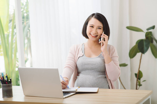 Genç neşeli hamile kadın telefonda konuşuyor. Ayrıca bilgisayarı kullanarak uzaktan çalışıyor. - Fotoğraf, Görsel