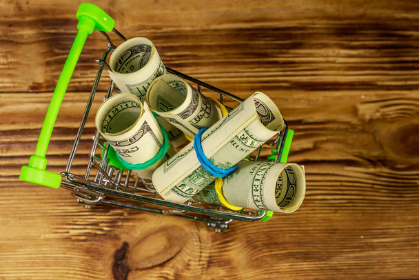 Small shopping cart with rolls of one hundred dollar bills on wooden background. Consuming concept - Photo, Image