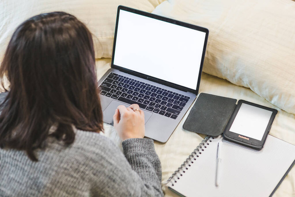 Женщина, работающая над ноутбуком, лежит в телефонной книге с видом на кровать. белый экран - Фото, изображение