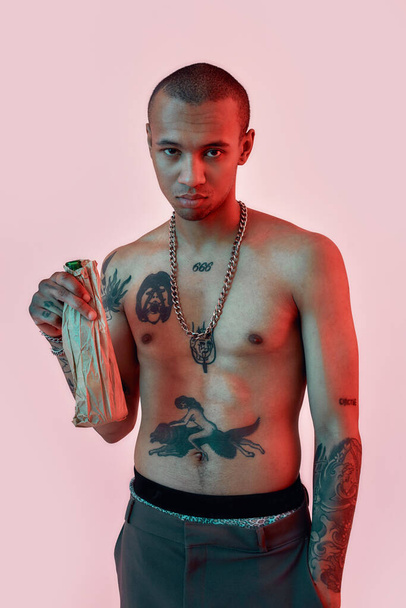 Drink responsibly. A dark-skinned male with tattoos standing topless holding a bottle in a paper bag - Фото, зображення