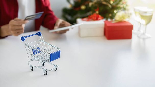 Online nákupní koncept. Nákupní košík na bílém stole s asijskou ženou držící smartphone a pomocí kreditní karty pro on-line nakupování a vánoční stromeček a dekorace pozadí. - Fotografie, Obrázek