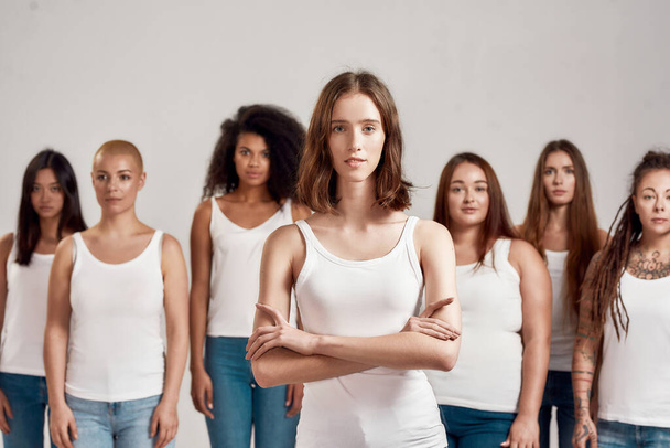 Portrait of young caucasian girl in white shirt looking at camera while standing with arms crossed. Group of diverse women posing isolated over grey background - Foto, Bild