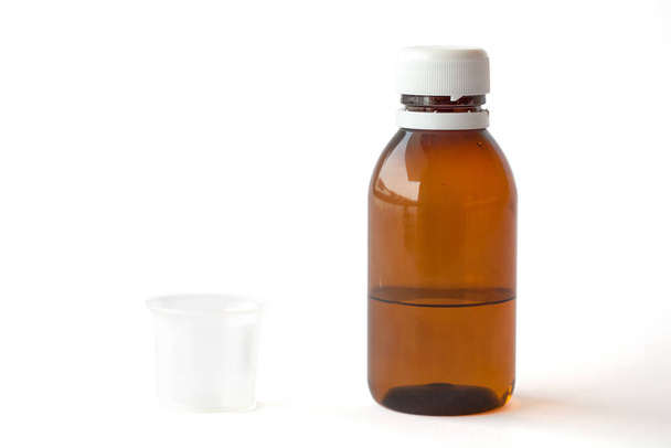 Διαφανές πλαστικό μπουκάλι με υγρό φάρμακο και ένα μικρό κύπελλο μέτρησης σε λευκό φόντο με μονοπάτια εργασίας - Φωτογραφία, εικόνα