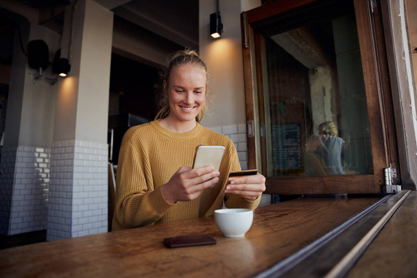 Mujer joven haciendo el pago en línea con tarjeta y teléfono inteligente mientras se sienta y disfruta del café en la cafetería - Foto, Imagen