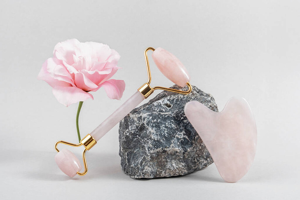 Crystal rose quartz facial roller and massage tool jade Gua sha on stones and pink flower on grey background. Facial anti-age massage for natural lifting and toning treatment at home. - Photo, Image