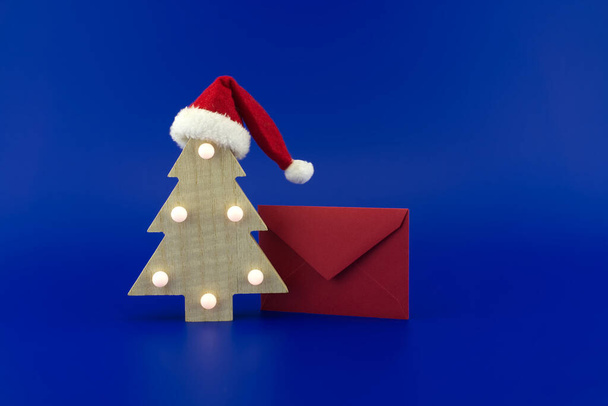 Stylizowana drewniana choinka w kapeluszu Mikołaja na górze obok czerwonej koperty na pozdrowienia na świątecznym niebieskim tle. Koncepcja nowego roku i świąt Bożego Narodzenia - Zdjęcie, obraz