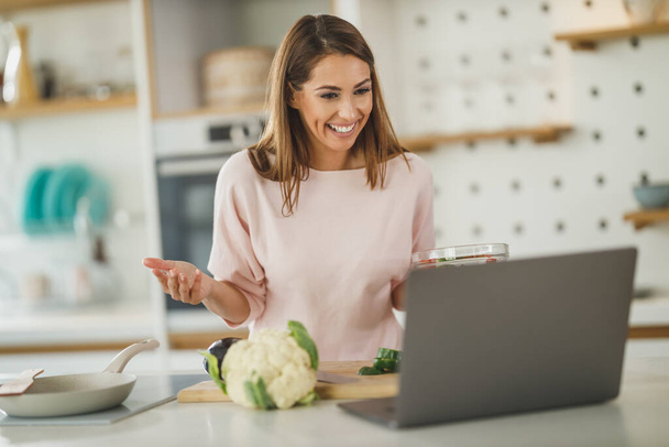 Inyección de una mujer joven usando un ordenador portátil para hacer un vlog con alguien mientras prepara una comida saludable en casa. - Foto, imagen